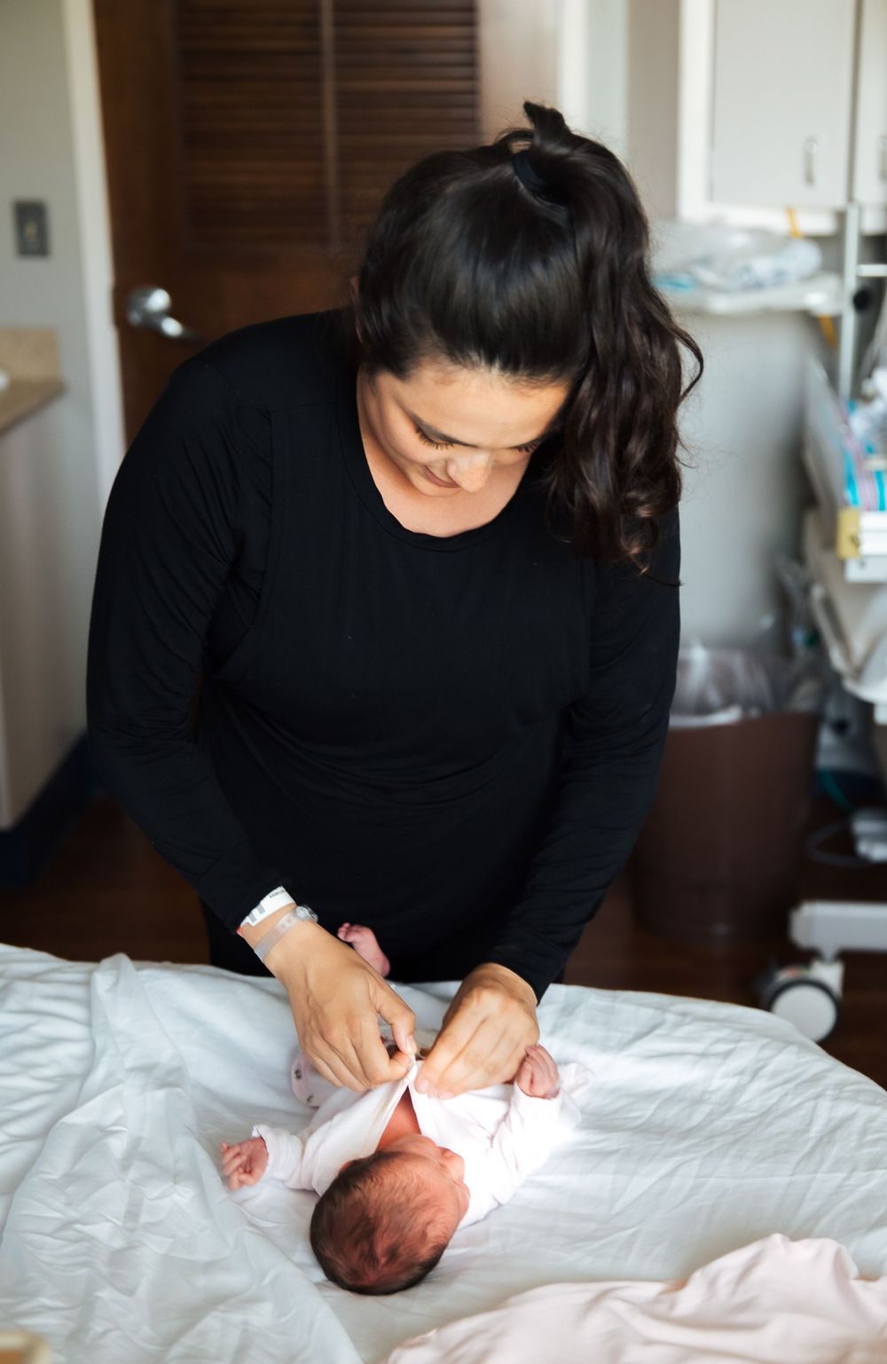 A mother in a black top dresses her newborn baby on a hospital bed after giving birth.  fresh 48, Newport Hospital