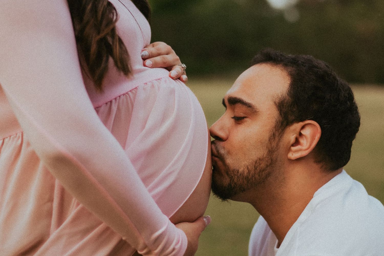 A man kisses the baby bump of his fiancé, who is dressed in a soft, pink maternity dress.