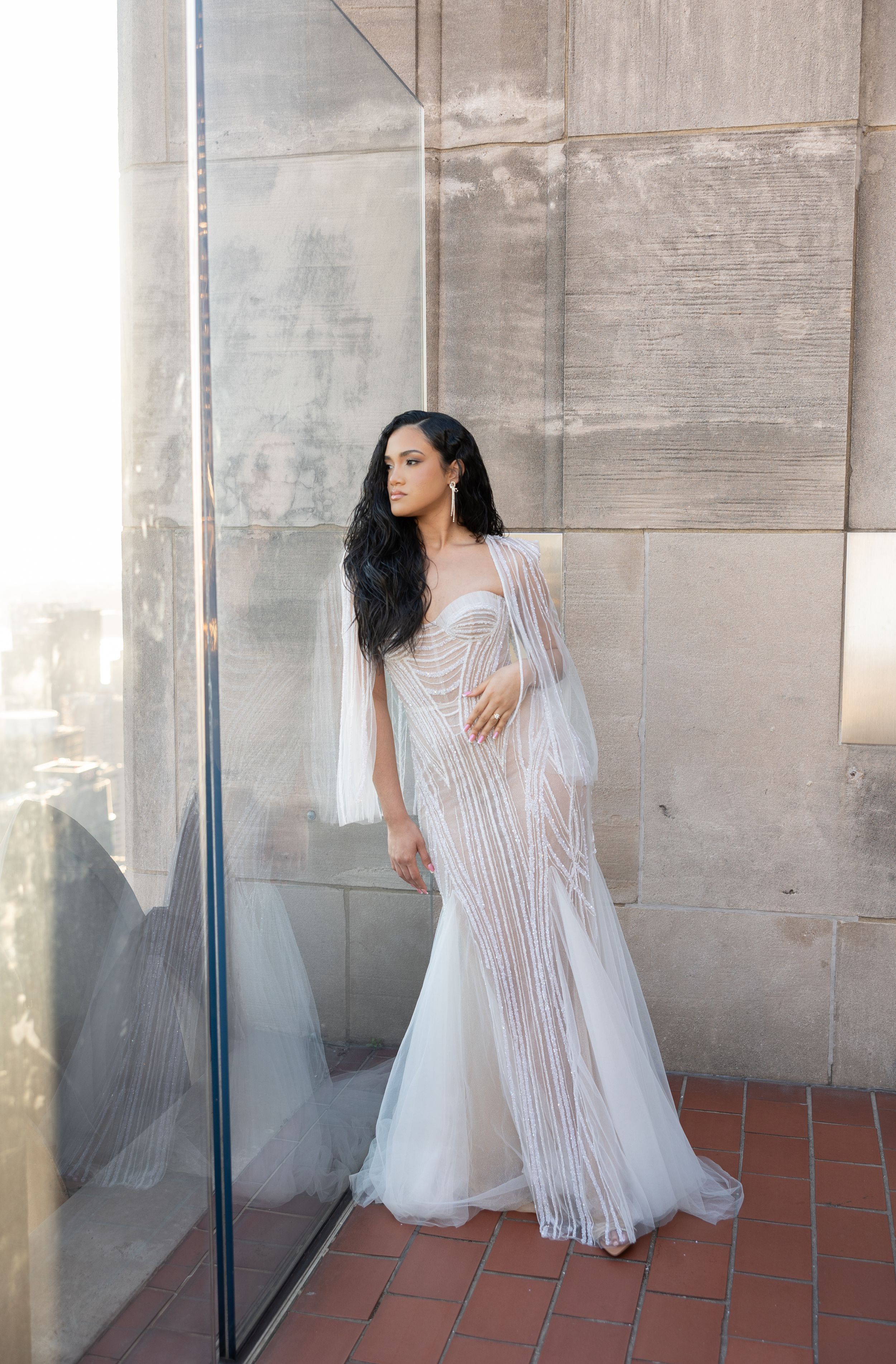 Woman in white beaded wedding gown poses against stone wall with dramatic lighting.
