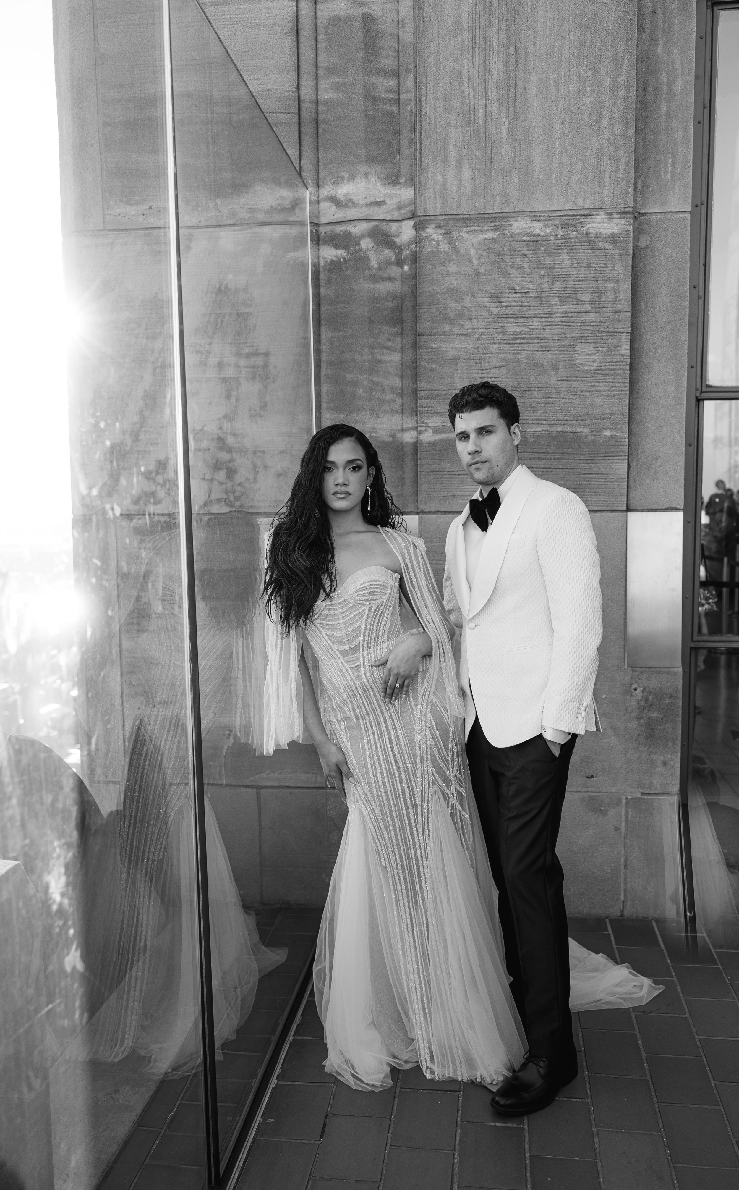 Couple in formal wear pose together against industrial backdrop in black and white.
