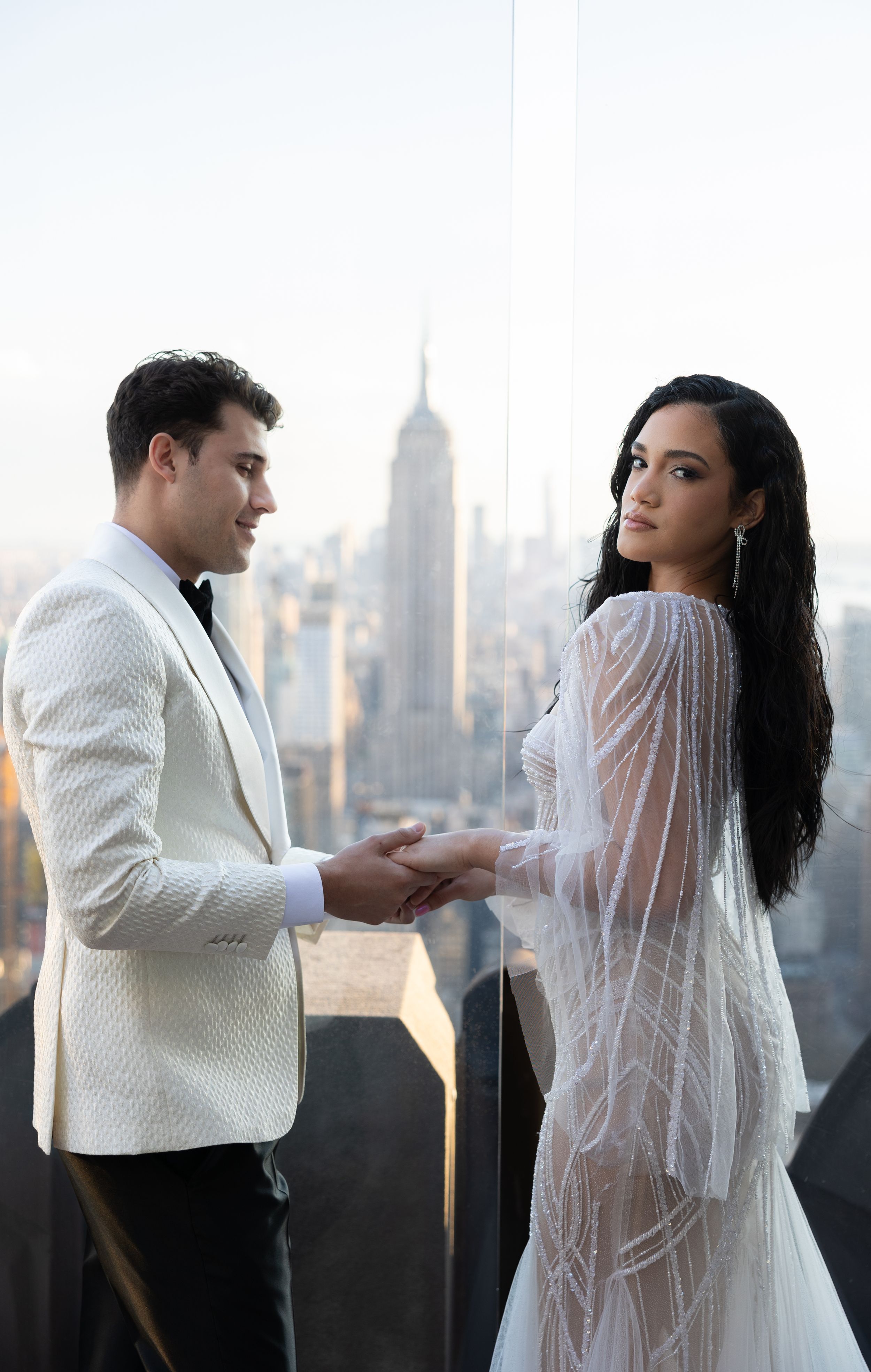 Couple holds hands in elegant wedding attire on NYC rooftop with iconic skyline in background.