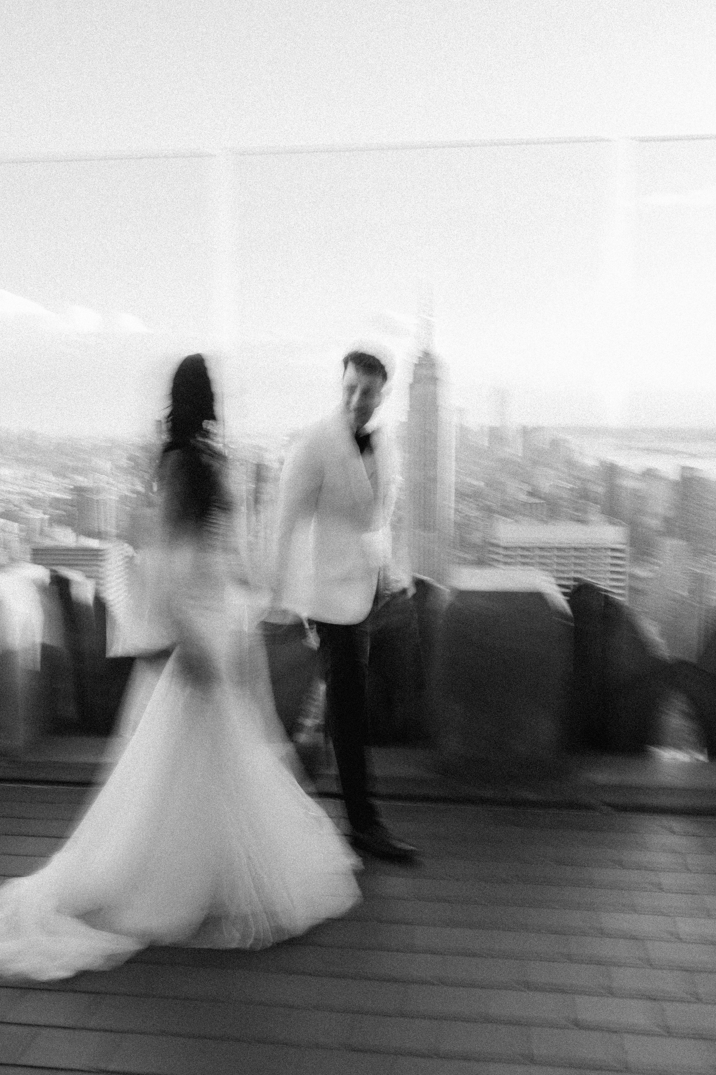 Artistic blurred black and white photo of couple in wedding attire on city rooftop.