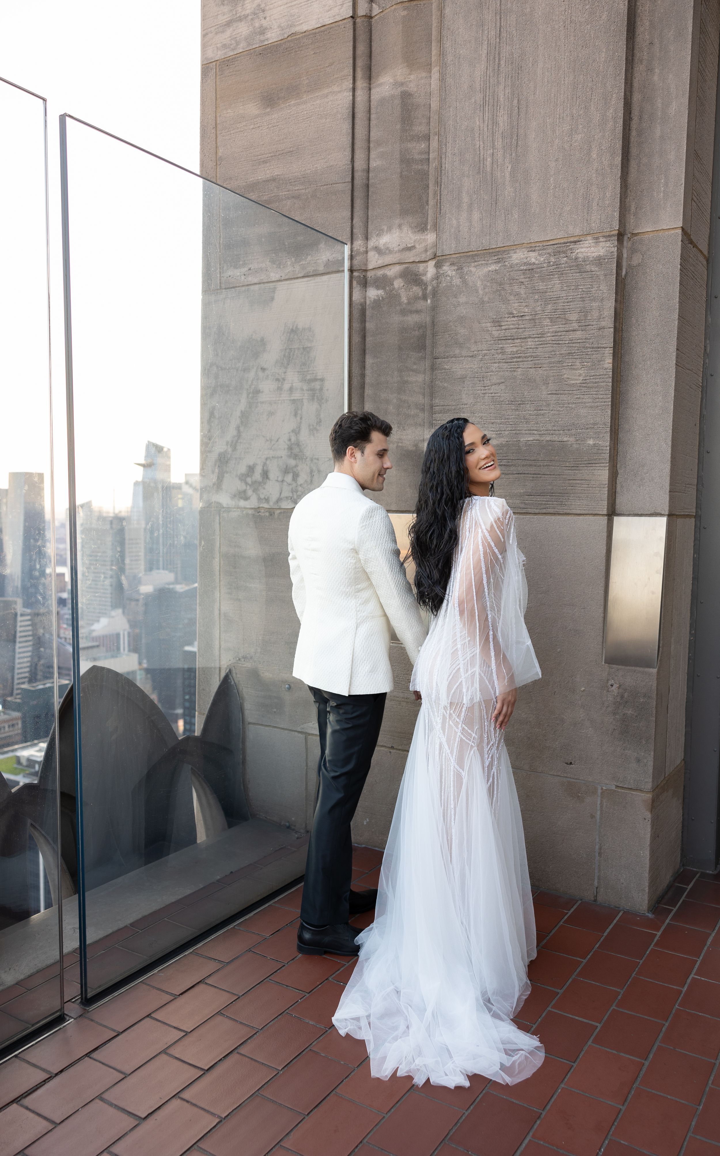 Couple in wedding attire standing against glass wall with NYC skyline view.