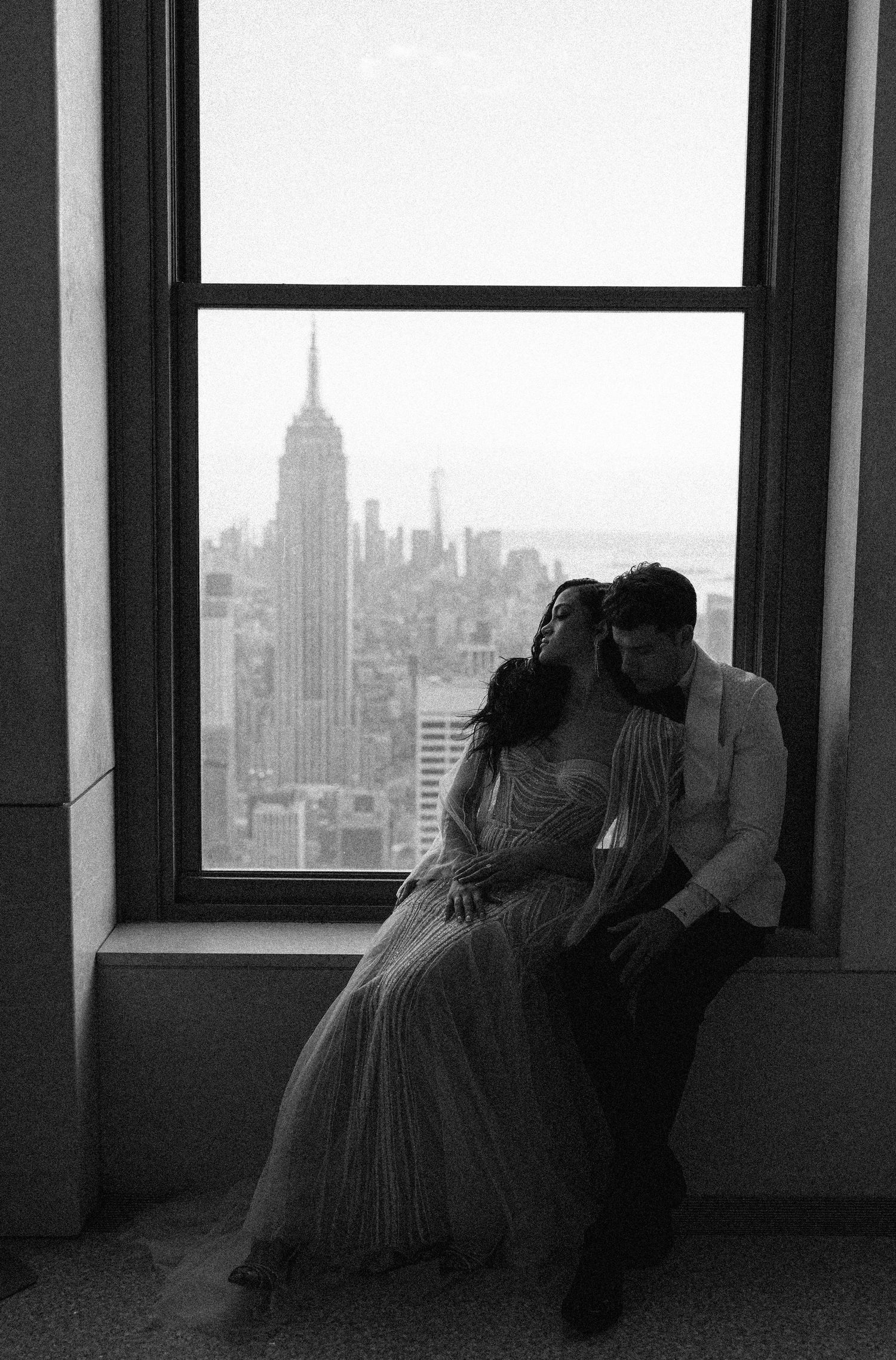 Romantic couple sitting by window overlooking Empire State Building in black and white photography.