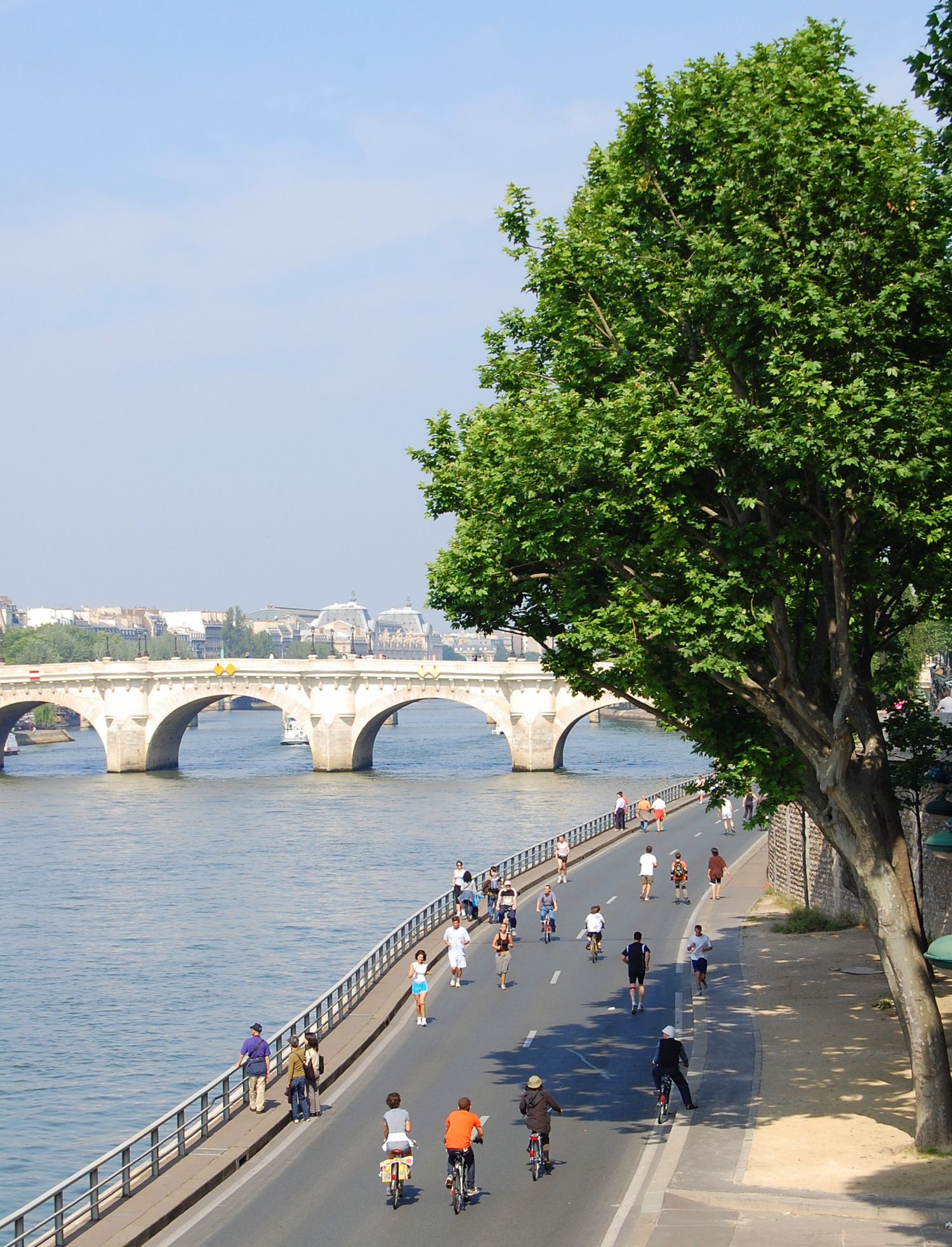 People cycling and walking along Seine River path under stone bridge in Paris.