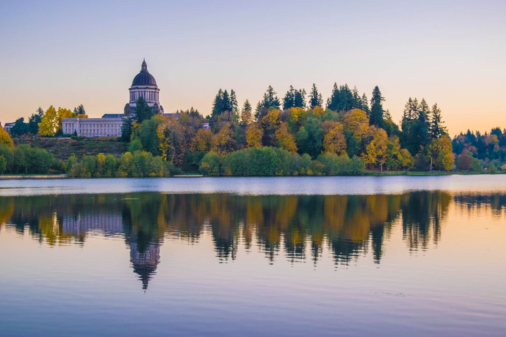 Capitol building reflects in calm lake water surrounded by autumn trees at sunset.