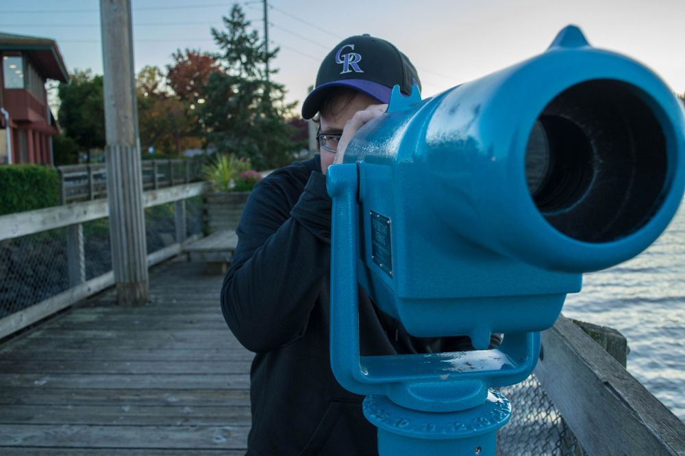 Someone looking through a large blue viewfinder telescope on a waterfront boardwalk.