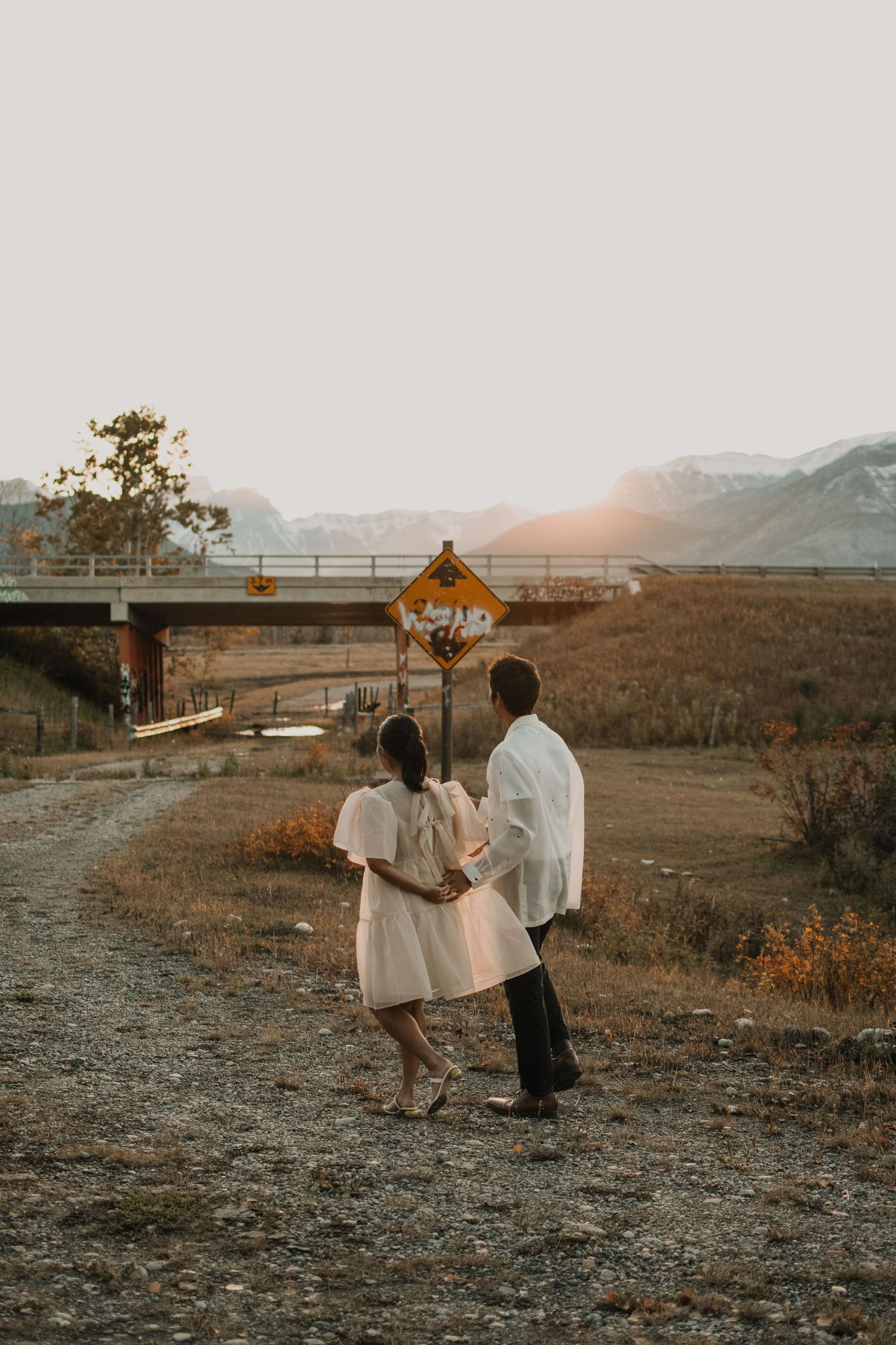 Couple walking together on dirt path during golden sunset near bridge.