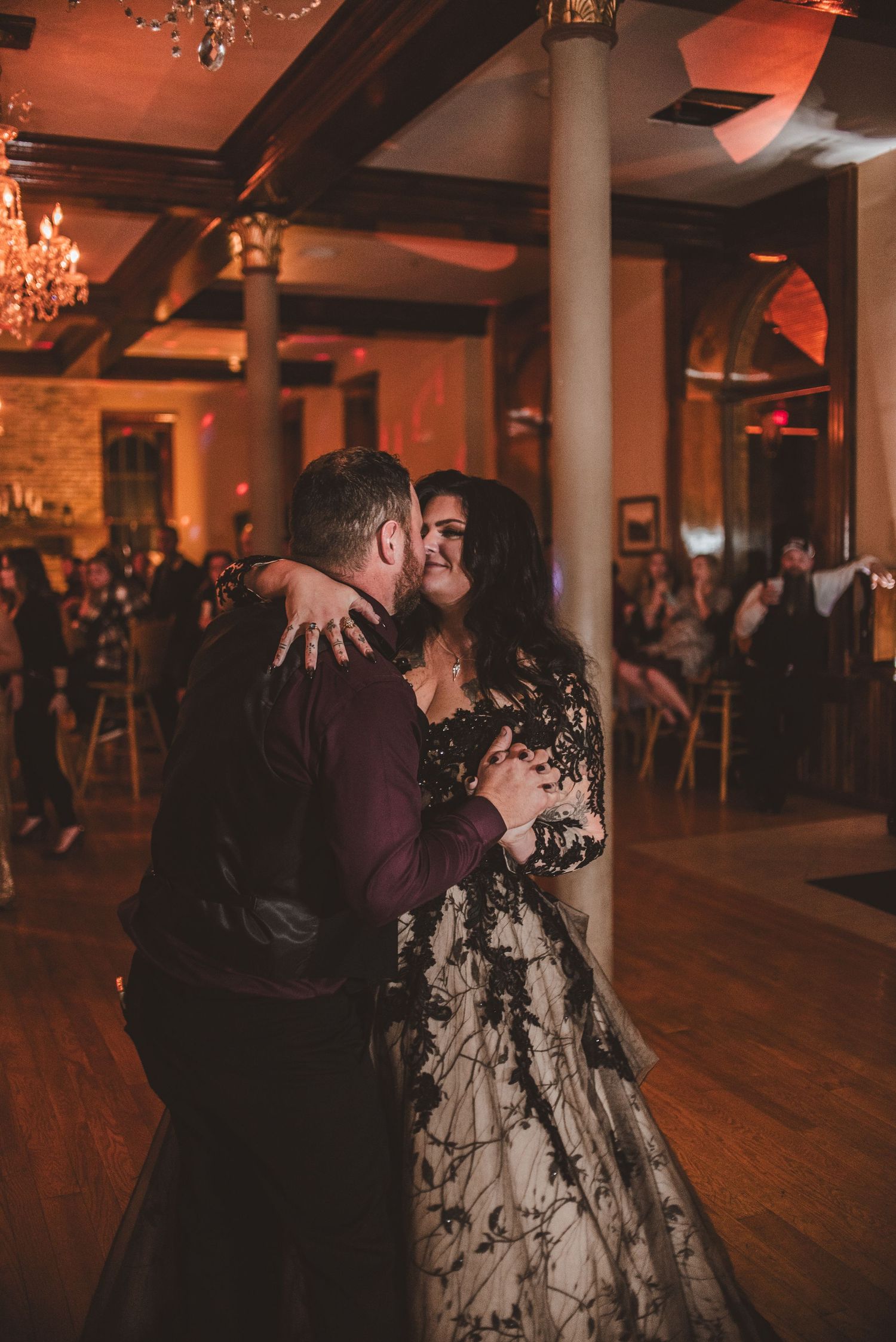 A couple dances intimately under warm lighting in an elegant ballroom with exposed brick walls and crystal chandeliers.