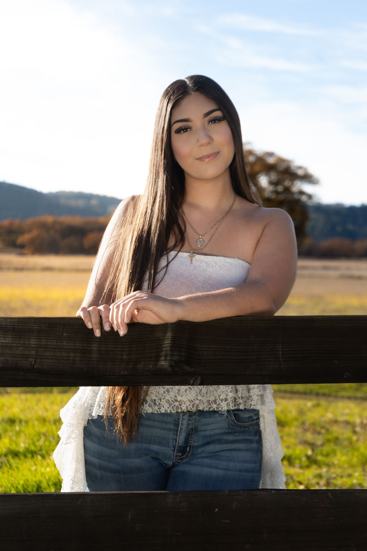 Young woman with long dark hair in white top and jeans leaning on wooden fence in sunny field.