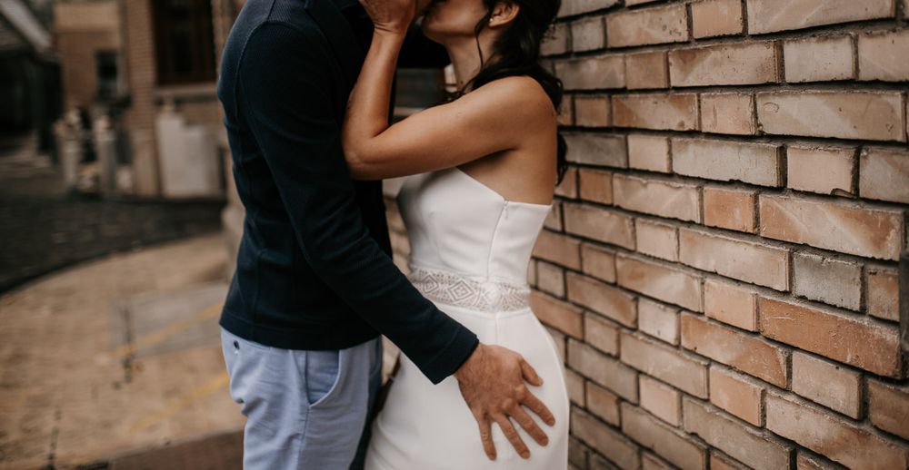 Groom embracing bride in white wedding dress against textured brick wall.