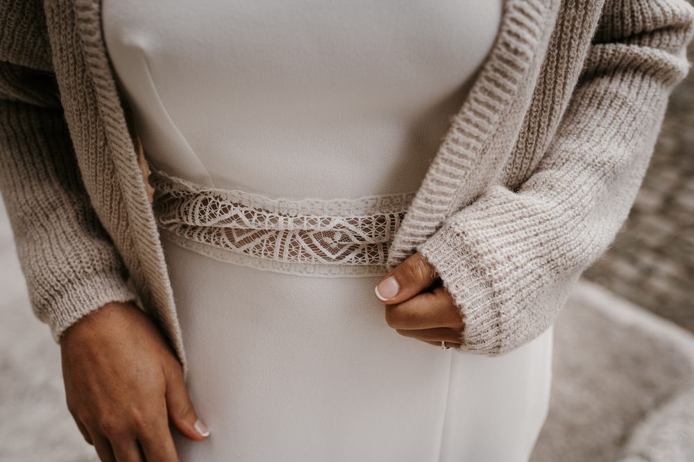 Close-up of bride wearing white wedding dress with lace belt and knit cardigan.