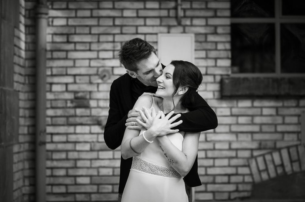 Black and white photo of groom embracing bride from behind near brick wall.