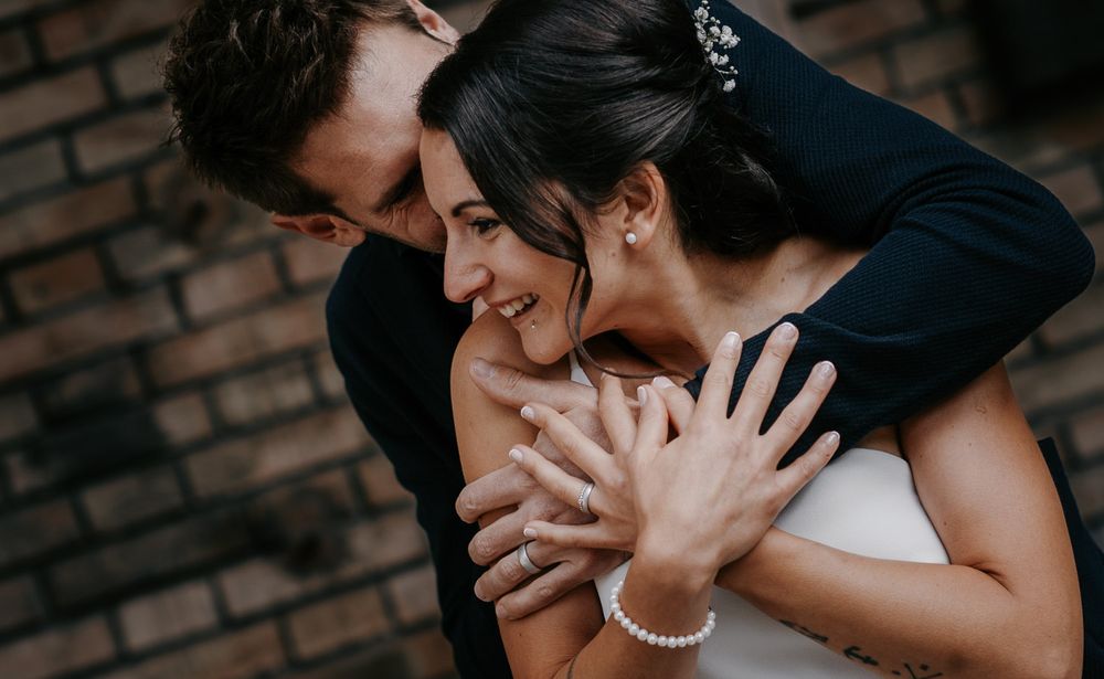 Groom embracing smiling bride from behind showing wedding rings near brick wall.