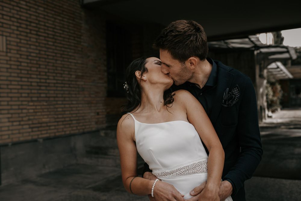 Couple shares romantic kiss against brick wall, bride in elegant white gown with delicate belt detail.