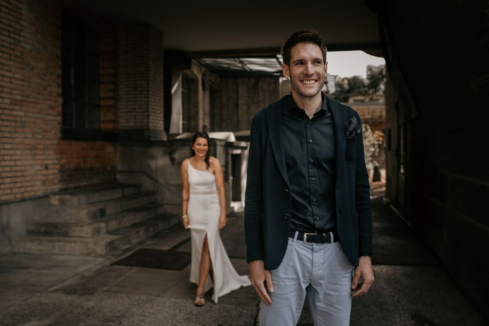 Smiling groom in black shirt stands in foreground as bride in white dress walks behind him.