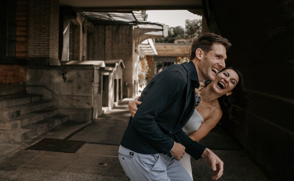 Laughing couple embraces playfully in rustic outdoor alleyway during casual engagement photography session.