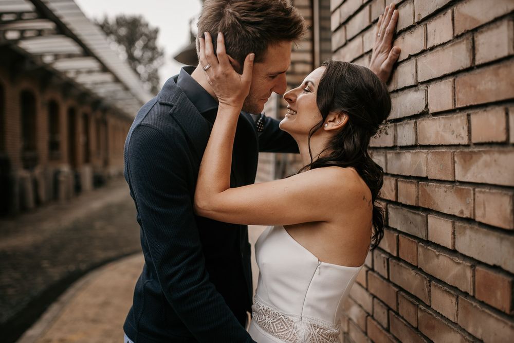 Joyful bride and groom sharing playful kiss against brick wall during engagement shoot.