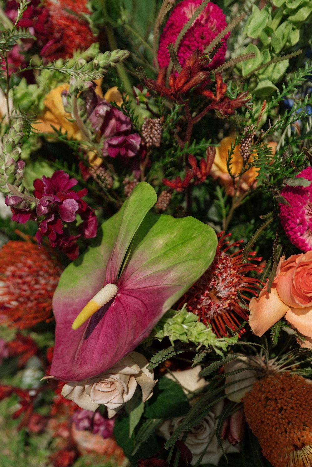 Vibrant tropical floral arrangement featuring anthurium, pincushion protea, roses, and lush greenery in rich jewel tones.