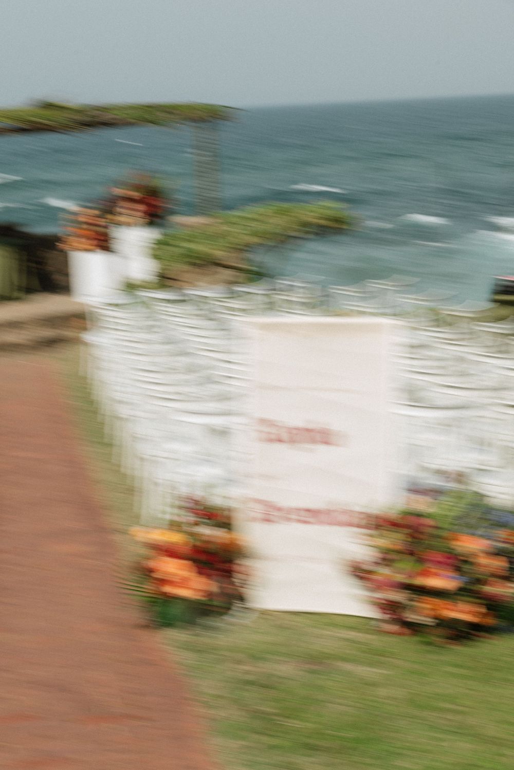 Blurry outdoor wedding ceremony setup with white draping and orange floral arrangements overlooking a turquoise ocean.