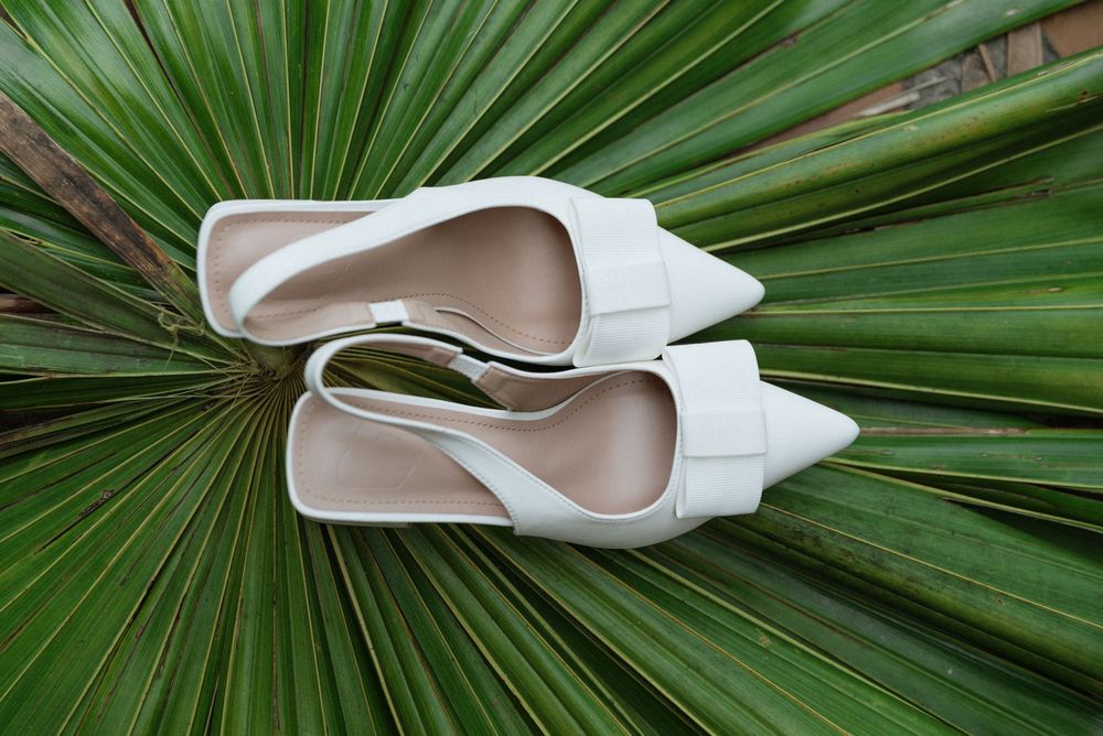 White pointed-toe slingback heels with bow detail displayed on a tropical palm leaf background.