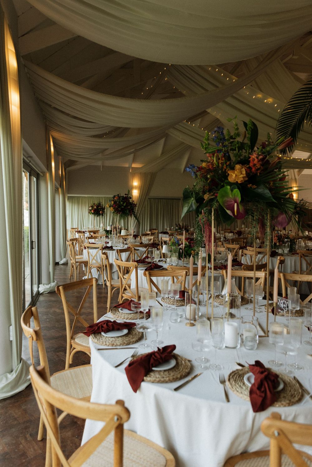 Elegant wedding reception with white draped ceiling, wooden cross-back chairs, wicker charger plates, burgundy napkins, and tall tropical fl