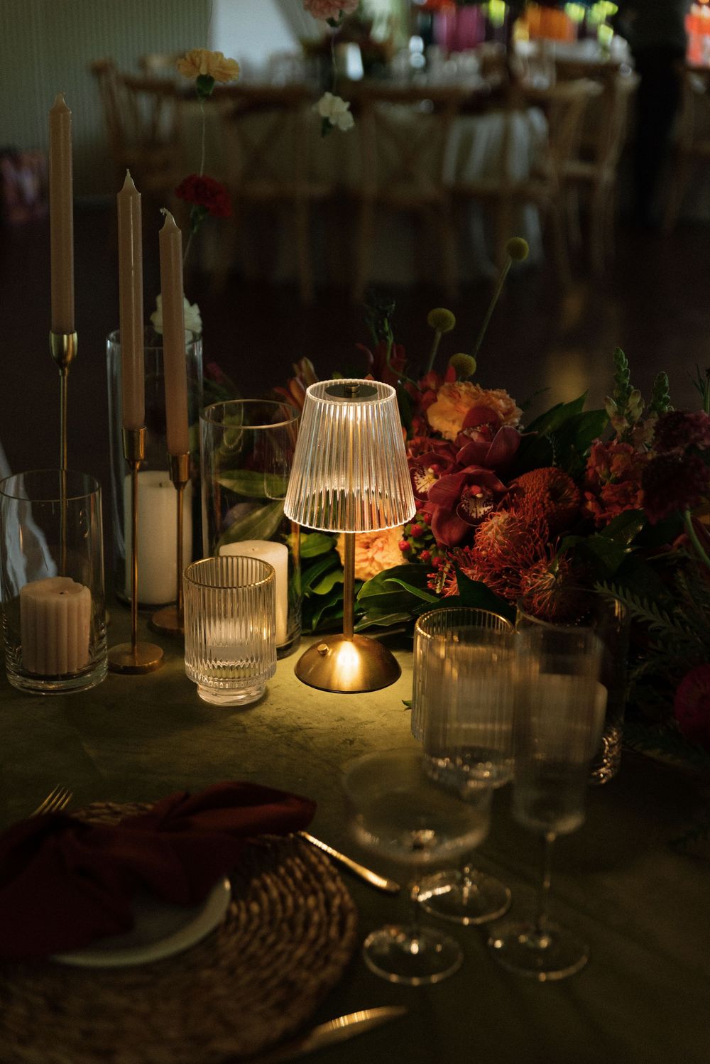 Elegant wedding table setting with glowing lamp, candles, crystal glasses, floral centerpiece, and burgundy napkins in dim light.