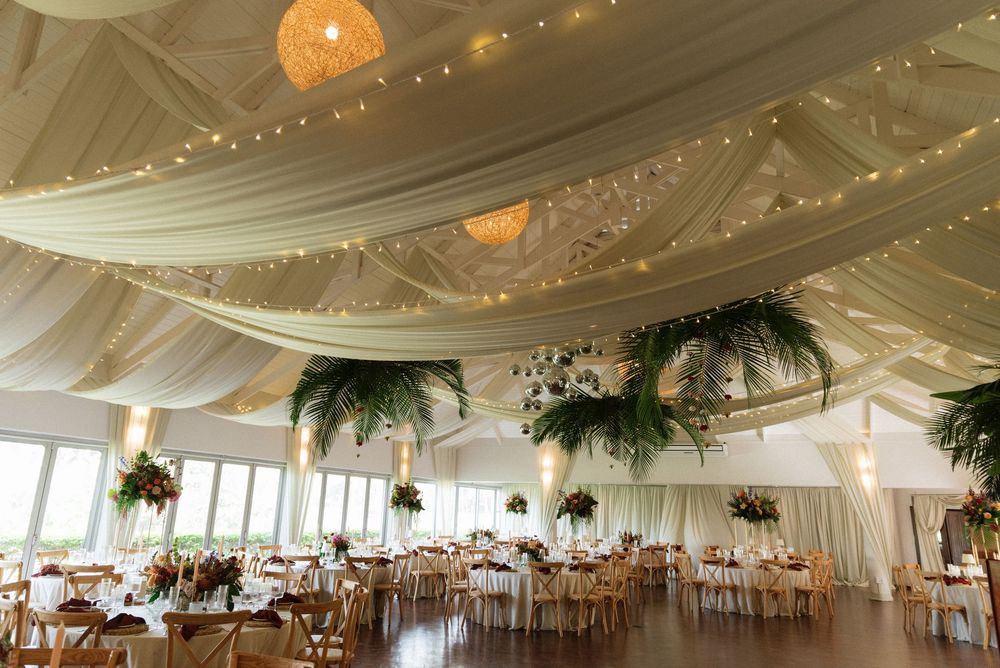 Elegant wedding reception hall with white draped ceiling, fairy lights, tropical greenery centerpieces, and wooden chairs at round tables.