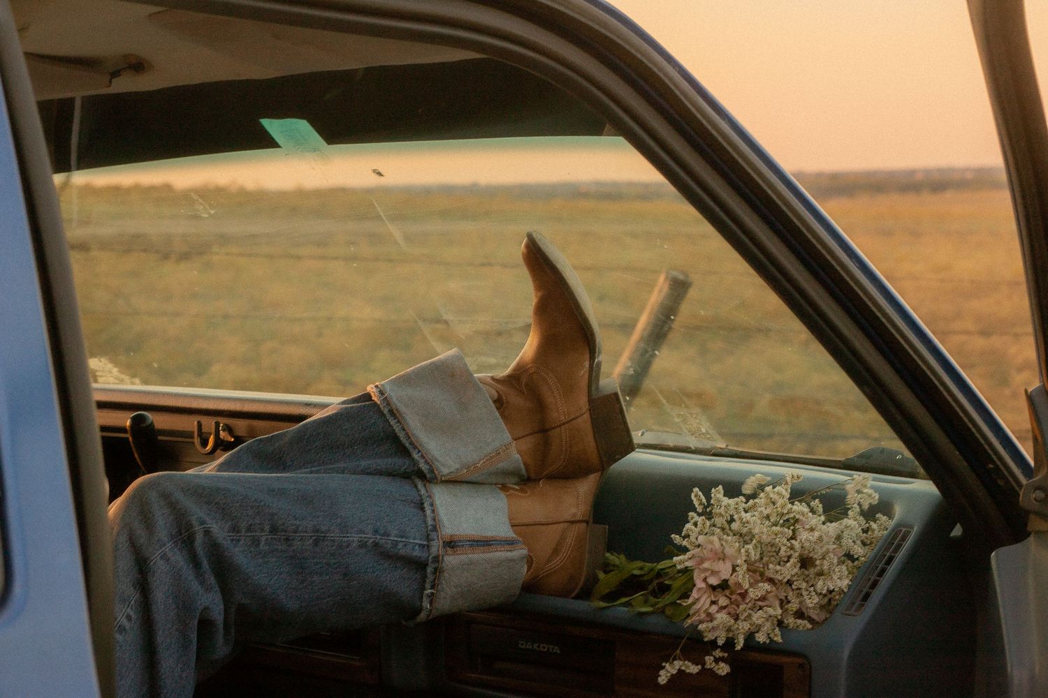 Person relaxing in vintage truck cab with cowboy boots up, wildflowers beside them, golden sunset over open fields.
