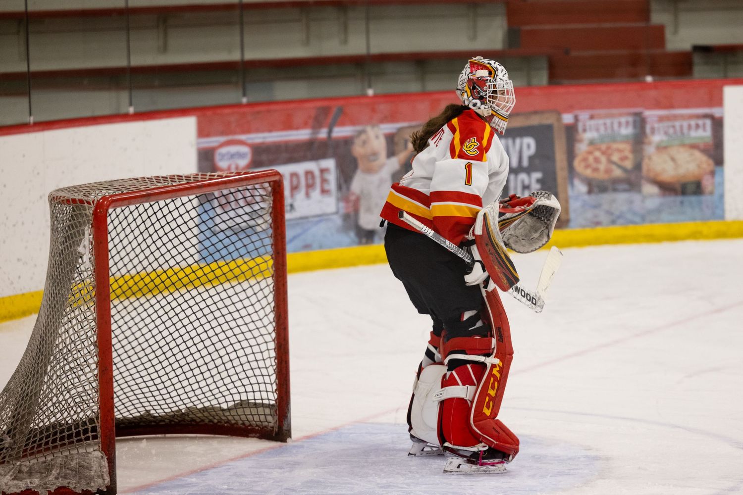 Ice hockey goalie in red and yellow uniform stands ready in front of net on indoor rink.