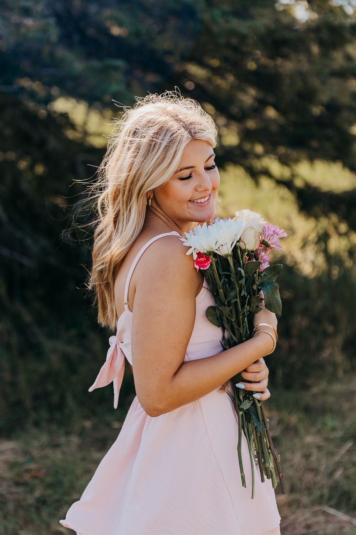 A person in a pink dress holds and smells a bouquet of fresh flowers in a sun-dappled outdoor setting.