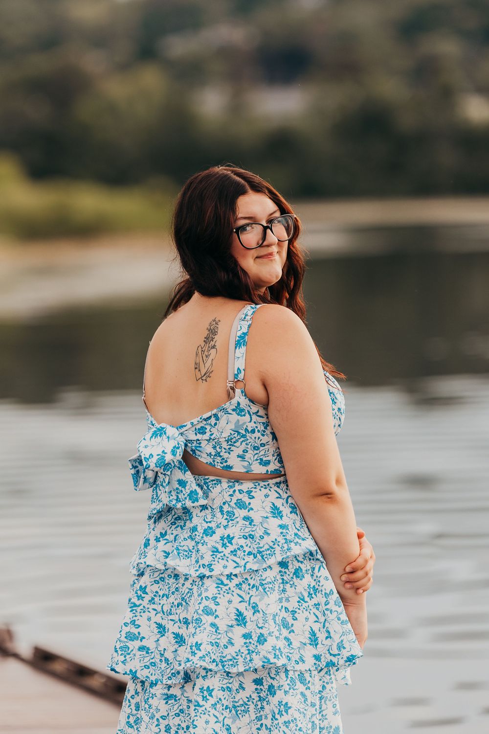 A person wearing a blue and white floral summer dress stands by a lake, looking over their shoulder with a smile.