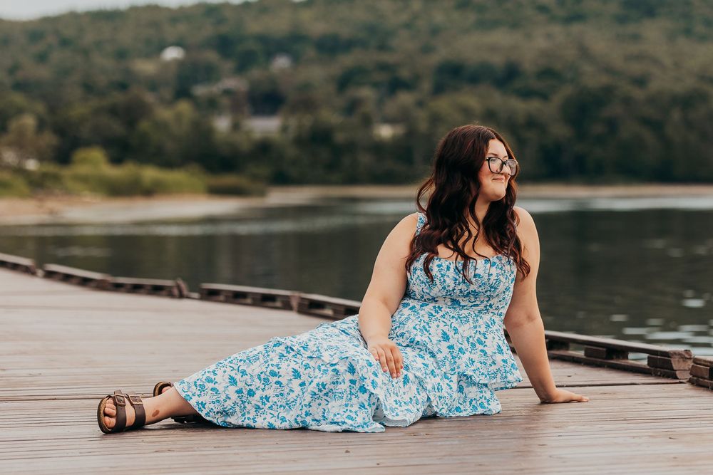 A person in a blue floral dress lies on a wooden dock by a serene lake during a summer afternoon.