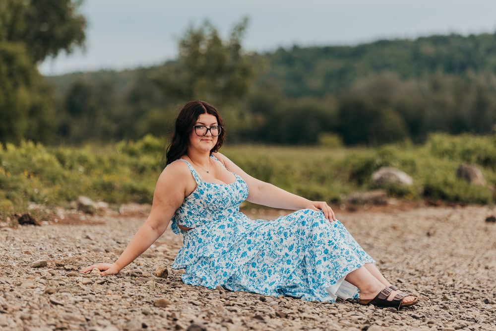 A person in a blue floral sundress sits on a rocky beach with green hills in the background on a sunny day.