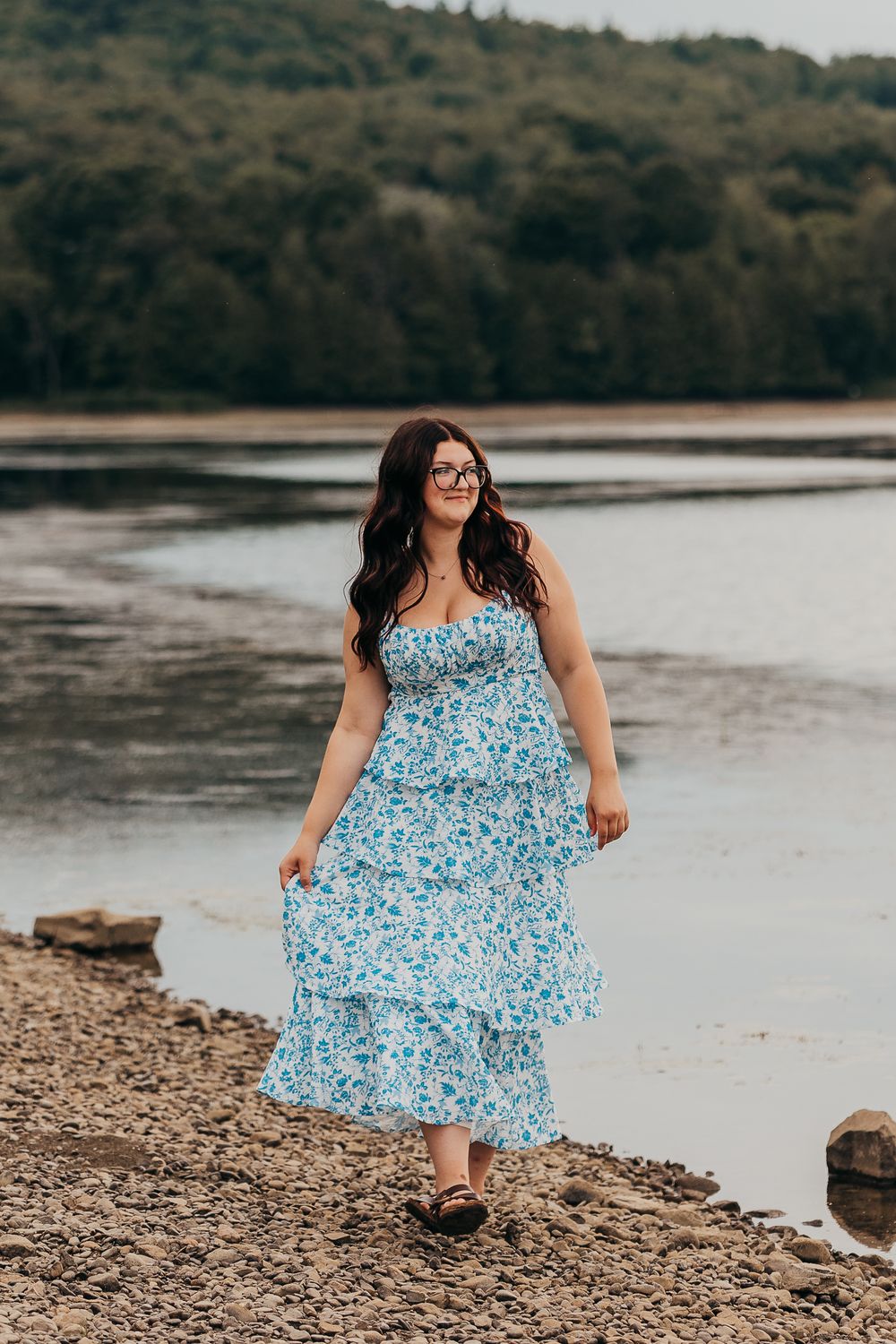A person in a flowing blue floral dress poses along a rocky lakeshore on an overcast day.