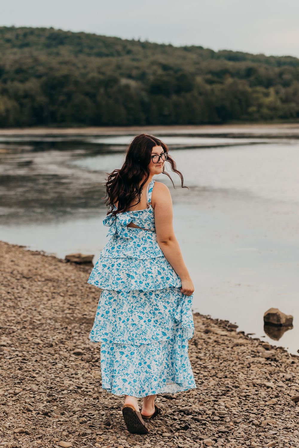 Woman in flowing blue floral tiered maxi dress stands on rocky shoreline by tranquil lake at dusk.