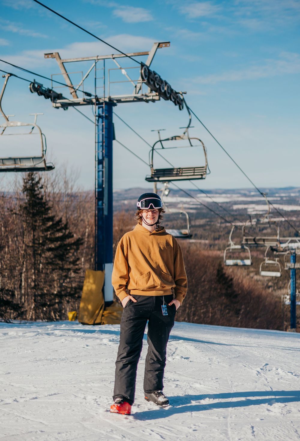 Skier in brown jacket glides down snowy slope with chairlift cables overhead on sunny winter day at ski resort.