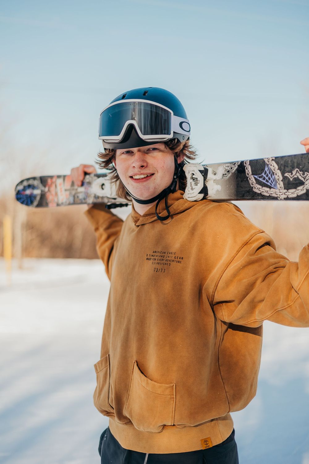 A snowboarder in a tan jacket rides across snowy terrain on a bright winter day.