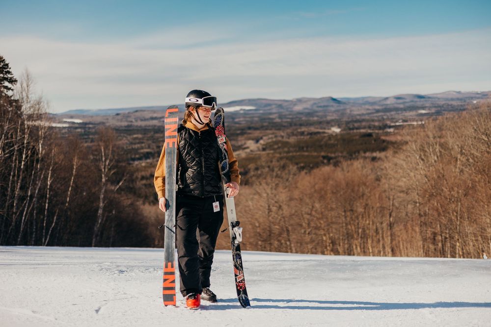 A skier stands on a snowy slope overlooking a mountainous winter landscape with bare trees and blue skies.