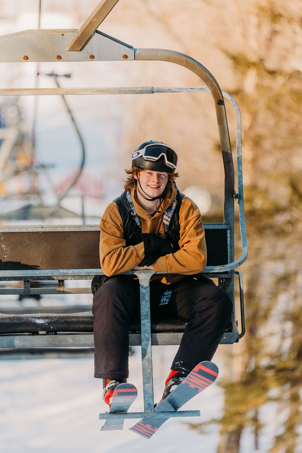 A skier in a brown jacket and ski gear sits on a chairlift against a winter mountain backdrop.