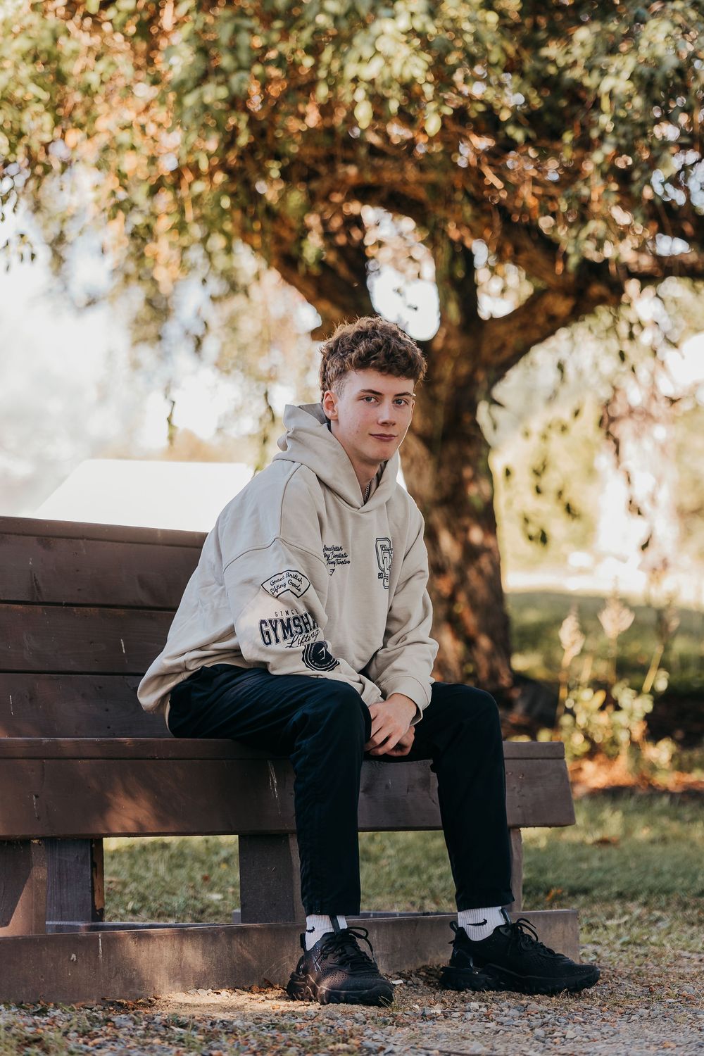 Person wearing a light-colored hoodie sits thoughtfully on a wooden bench under autumn trees in a park setting.
