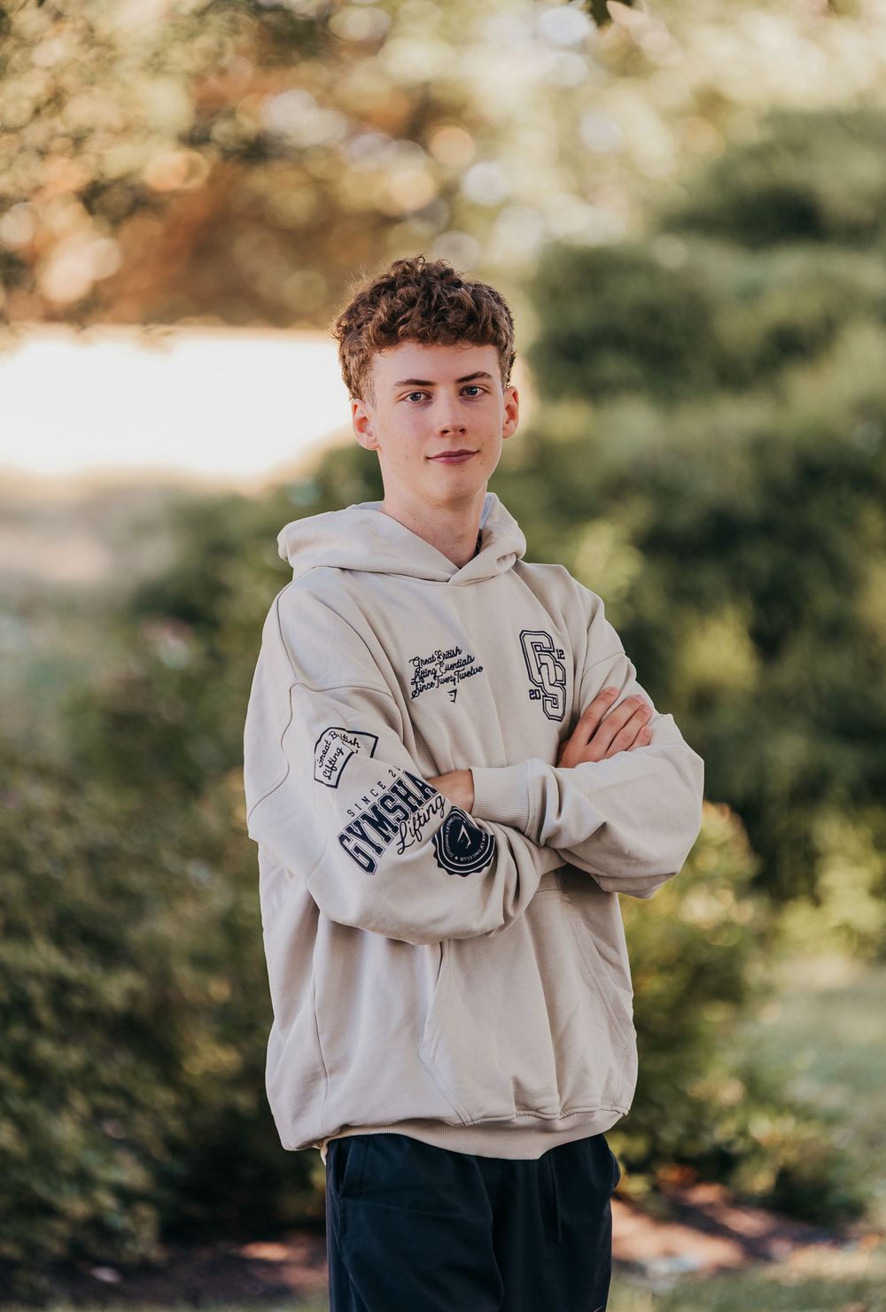 A young person in a white oversized hoodie poses outdoors with arms crossed against a blurred natural background.
