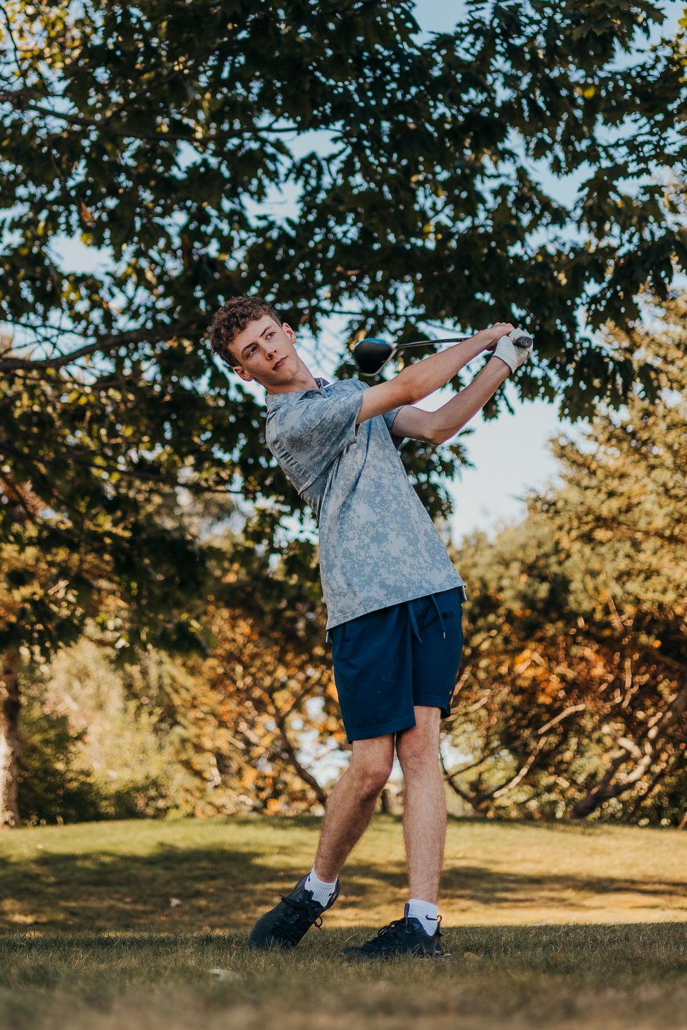 A golfer in a gray t-shirt and navy shorts practices their swing form on a sunny day with trees in the background.