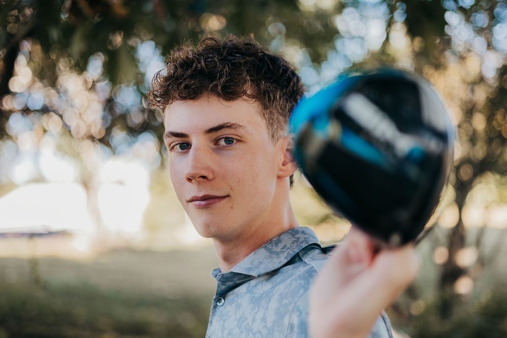 Someone holds up a black motorcycle helmet against a blurred outdoor background with trees and sunlight.