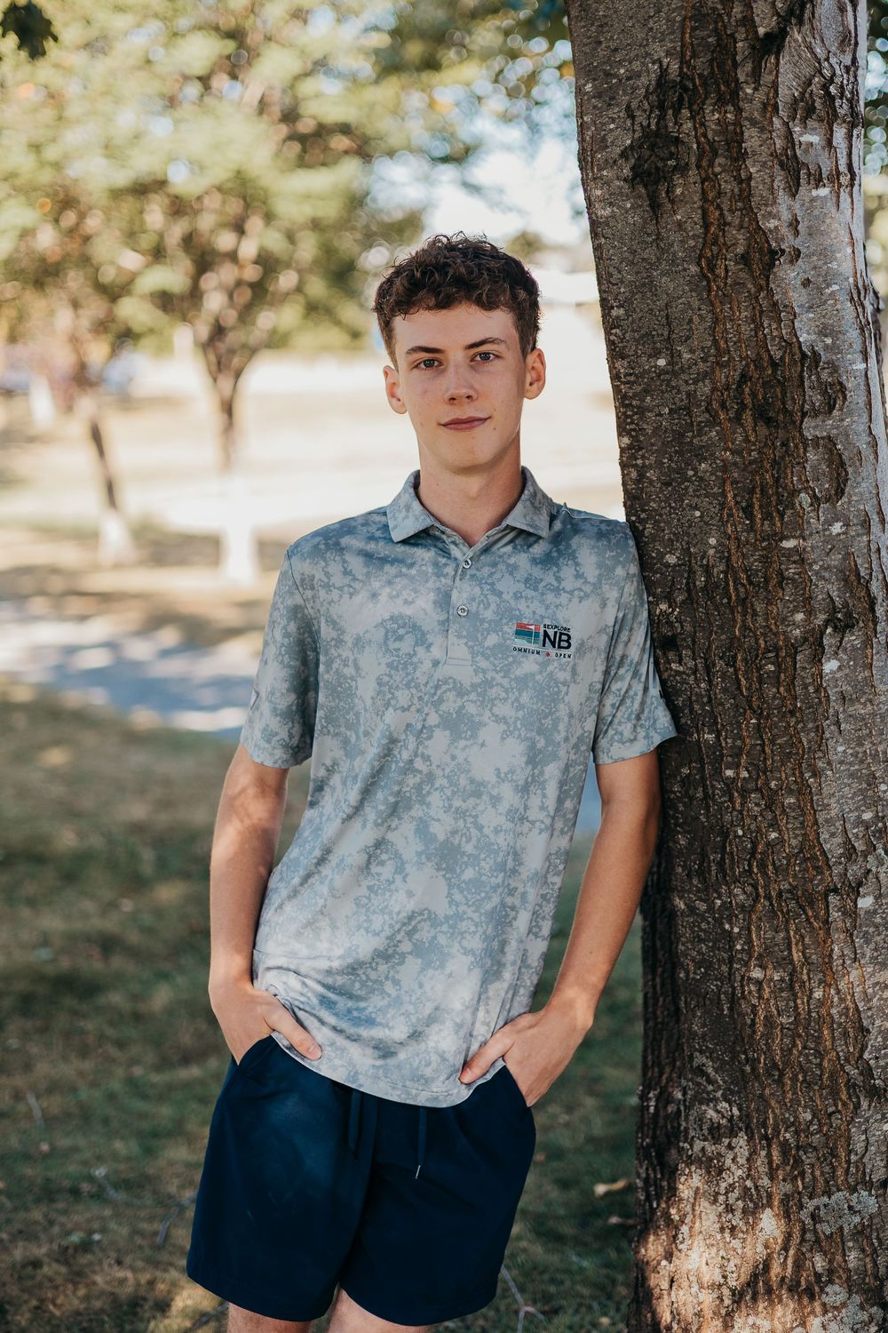 A person in a light blue tie-dye polo shirt and navy shorts stands casually next to a tree trunk in a sunny park setting.