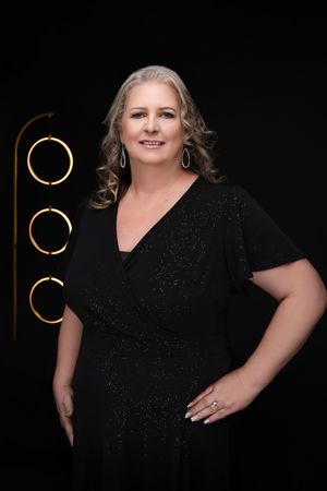 Professional portrait against black background shows person in sparkly black dress with silver curly hair and dangling earrings.