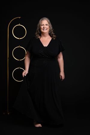 Woman in black dress poses elegantly against dark background with gold circular decorative elements.