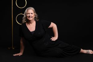 A person in a black dress poses elegantly on a dark floor against a black background with gold circular decor.