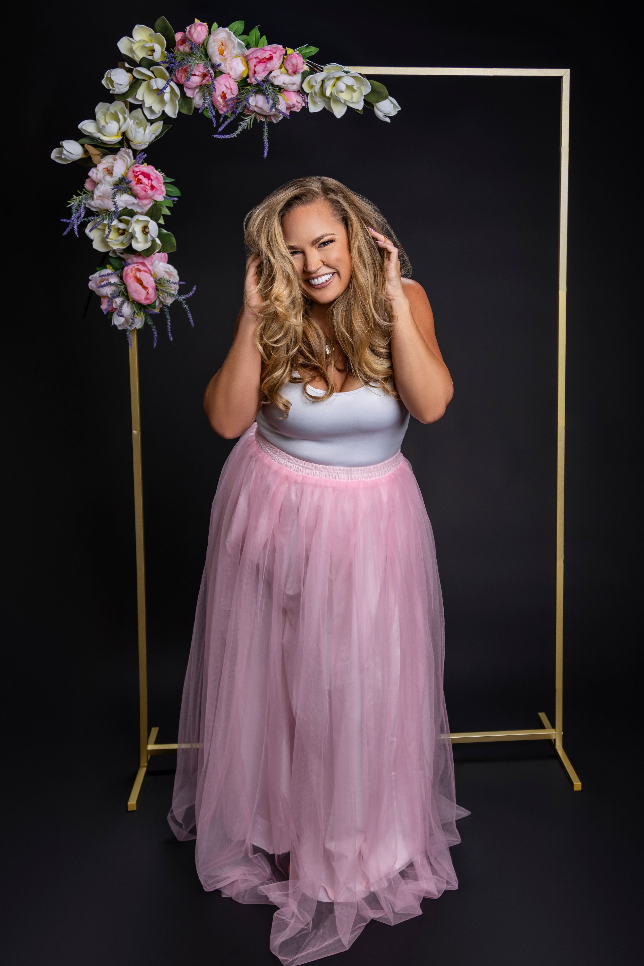 A romantic studio portrait with a gold frame arch decorated with pink and white flowers and flowing pink tulle gown.