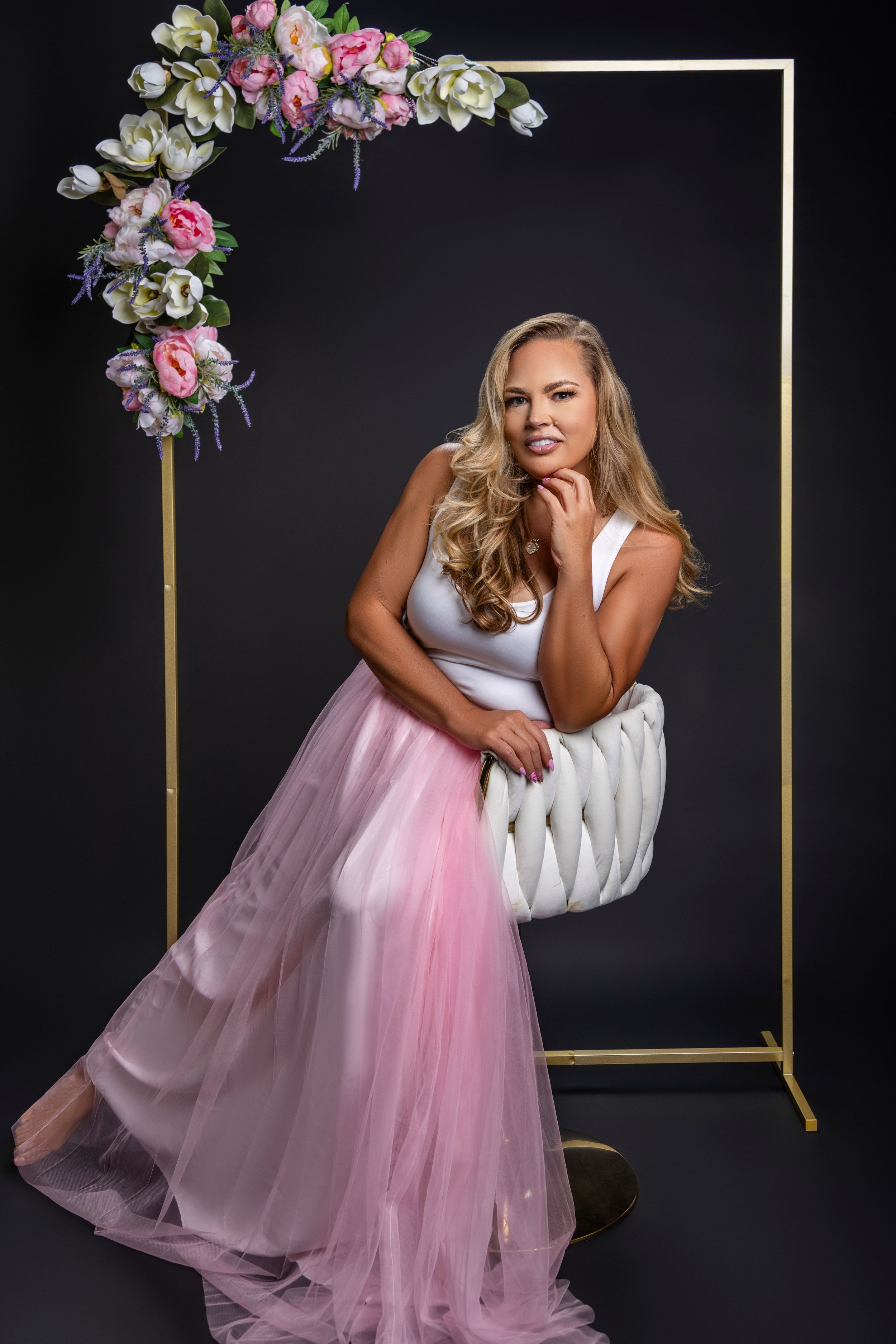 Elegant fashion portrait with pink tulle skirt and floral arch decoration against dark backdrop.